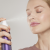 Na zdjęciu kobieta spryskuje twarz nawilżającą mgiełką Lavender Hydration Spray od Jane Iredale. Ma zamknięte oczy i zrelaksowany wyraz twarzy. Produkt widoczny w jej dłoni to czerwona butelka z białym atomizerem – idealny wrażliwych skór.