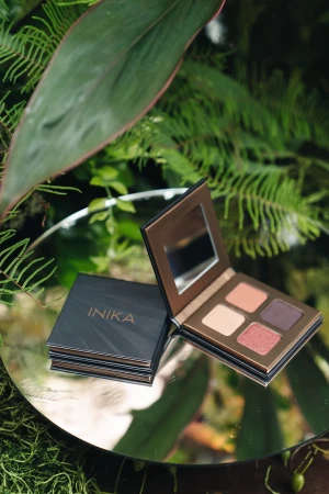 Na zdjęciu znajduje się paleta cieni do powiek marki INIKA Organic, ustawiona na okrągłym lustrze położonym wśród zielonych roślin i liści. Paleta jest otwarta, ukazując cztery odcienie cieni – dwa jasne i dwa ciemniejsze, w ciepłej tonacji.