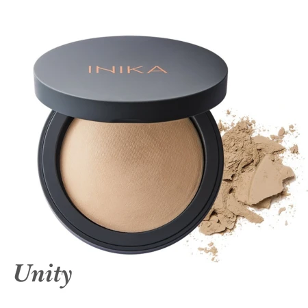 Na zdjęciu widoczny jest puder mineralny INIKA Organic w odcieniu Unity. Produkt znajduje się w eleganckim, okrągłym opakowaniu z ciemnym wieczkiem, na którym widnieje logo INIKA w złotym kolorze. Obok pudru widać rozsypaną niewielką ilość produktu