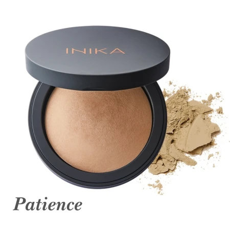 Na zdjęciu widoczny jest puder mineralny INIKA Organic w odcieniu Patience. Produkt znajduje się w eleganckim, ciemnym okrągłym opakowaniu z wieczkiem ozdobionym logo INIKA w złotym kolorze. Obok opakowania rozsypana jest niewielka ilość pudru