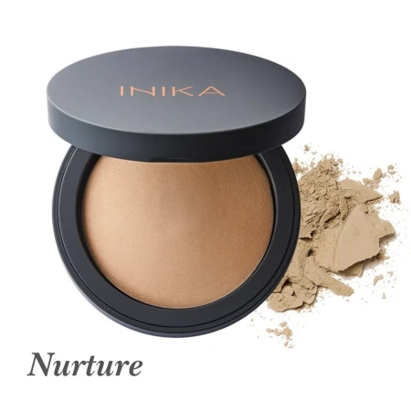 Na zdjęciu widoczny jest puder mineralny INIKA Organic w odcieniu Nurture. Produkt znajduje się w okrągłym, eleganckim opakowaniu z ciemnym wieczkiem i logo INIKA w odcieniu miedziano-złotym. Obok opakowania rozsypana jest niewielka ilość pudru