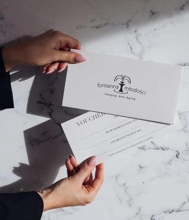 Elegancki voucher prezentowy z logo „Fontanna Młodości Instytut Anti Aging”, trzymany przez kobietę o zadbanych dłoniach i różowym manicure. Voucher znajduje się w dopasowanej kopercie.