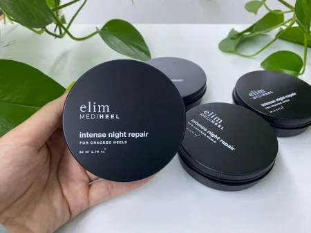 Na zdjęciu dłoń trzyma czarny pojemnik kremu elim MEDIHEEL Intense Night Repair na popękane pięty. W tle kilka identycznych pudełek na białym blacie i zielone liście rośliny, nadające naturalny charakter scenerii.
