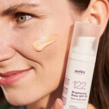 Widzimy kobietę, która trzyma w dłoni produkt Purles 122 Brightening Base SPF 50+. Na jej policzku nałożona jest porcja kosmetyku w odcieniu beżowym, co pokazuje konsystencję i kolor bazy. Skóra kobiety wygląda promiennie i zdrowo.