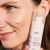Widzimy kobietę, która trzyma w dłoni produkt Purles 122 Brightening Base SPF 50+. Na jej policzku nałożona jest porcja kosmetyku w odcieniu beżowym, co pokazuje konsystencję i kolor bazy. Skóra kobiety wygląda promiennie i zdrowo.
