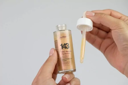 Na zdjęciu kobiece dłonie trzymahą butelkę serum Purles 143 VitC Serum Perfector w lewej dłoni. Druga dłoń trzyma pipetę, którą wyjęto z buteleczki – w pipetce znajduje się odrobina płynnego, lekko pomarańczowego serum.