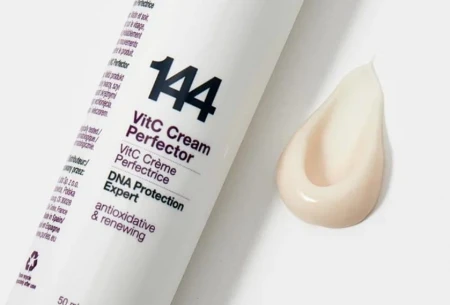 Na zdjęciu widoczna jest butelka kremu Purles 144 VitC Cream Perfector w białym, smukłym opakowaniu z fioletowymi i czarnymi napisami. Obok opakowania znajduje się próbka produktu – krem w odcieniu jasnego beżu, o kremowej, gładkiej konsystencji.