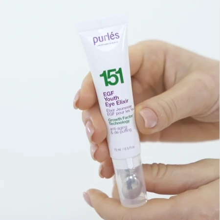 Na zdjęciu widać rękę trzymającą kosmetyk Purles EGF Youth Eye Elixir 151. To biały, smukły pojemnik w formie tubki o pojemności 15 ml, zakończony metalową kulką masującą, która ułatwia aplikację produktu i działa chłodząco na okolice oczu.