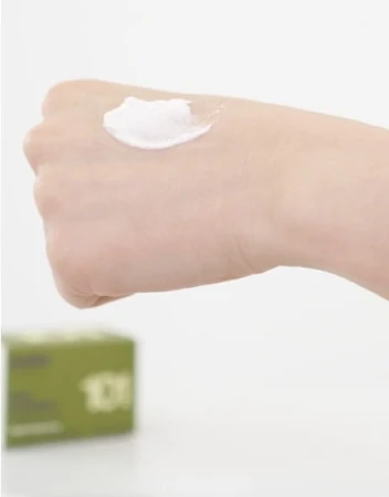 Na zdjęciu widać dłoń, na której rozprowadzono porcję kosmetyku Purlés Rice Exfoliator 101. Produkt ma kremową, gęstą konsystencję z delikatnym połyskiem. W tle, lekko rozmyte, widoczne jest opakowanie peelingu z numerem „101”.