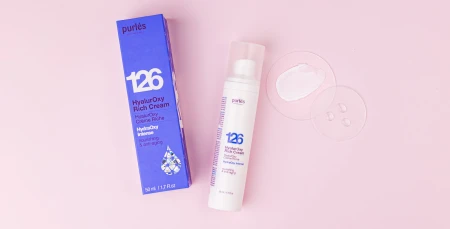Na zdjęciu jest krem HyalurOxy Rich Cream 126 w białym, smukłym opakowaniu z pompką, obok którego znajduje się jego kartonik. Obok produktu widoczna jest przezroczysta płytka z próbką kremu, co pozwala zobaczyć jego białą, kremową konsystencję.