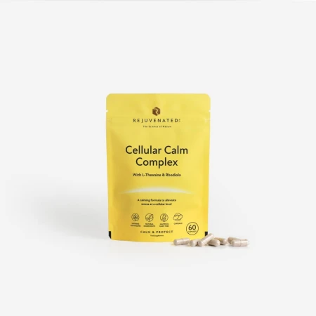 Zdjęcie przedstawia stojące, żółto-beżowe opakowanie suplementu diety w kapsułkach Rejuvenated Cellular Calm Complex. Obok opakowania, na gładkim, jasnoszarym tle, rozsypanych jest kilka beżowych kapsułek.