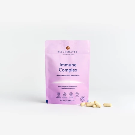 Zdjęcie przedstawia stojące, jasnofioletowe opakowanie suplementu diety w kapsułkach Rejuvenated Immune Complex.  Obok po prawej stronie leżą beżowe kapsułki