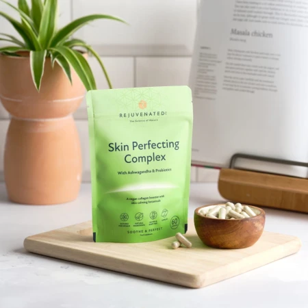 Zdjęcie przedstawia jasnozielone opakowanie suplementu Rejuvenated Skin Perfecting Complex, kapsułki w drewnianej misce, na drewnianej desce. W tle roślina doniczkowa i książka kucharska.