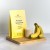 Myślę Szukam Zdjęcie przedstawia stojące, beżowe opakowanie proszku Rejuvenated Protein Smoothie o smaku bananowym. Na pierwszym planie, obok opakowania, znajduje się przezroczysta szklanka wypełniona kremowym napojem oraz kiść świeżych bananów