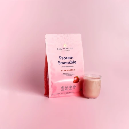 na jasnoróżowym tle stoi opakowanie produktu o nazwie "rejuvenated protein smoothie - strawberry. Jest to różowo-białe, stojące opakowanie w formie saszetki. Obok po prawej stronie, znajduje sie szklanka z jansoróżowym napojem