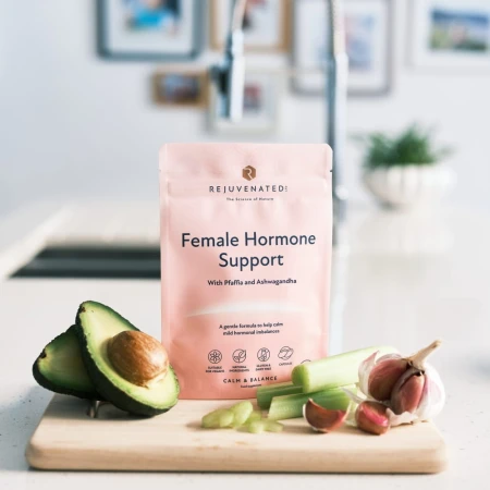 Na kuchennym blacie stoi różowa saszetka suplementu Rejuvenated Female Hormone Support z napisem „With Pfaffia and Ashwagandha”. Przed opakowaniem leży deska do krojenia z przeciętym awokado, łodygą selera naciowego i główką czosnku.