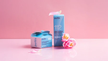 Na różowym tle ustawione są dwa niebieskie kosmetyki marki Purlés: po lewej sześcienne opakowanie produktu 149 Peptide Hydrogel Eye Patches z ozdobnym zamknięciem, a po prawej prostokątne pudełko kremu pod oczy 112 Age Control Eye Cream.