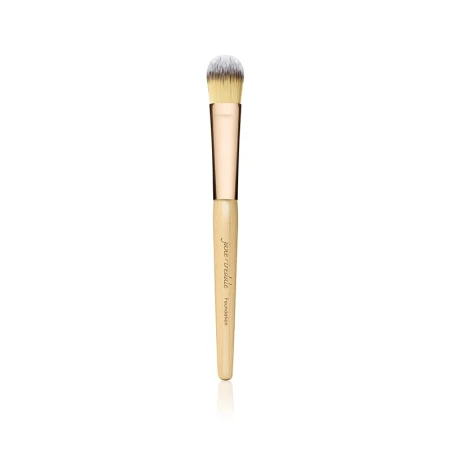 Na zdjęciu znajduje się pędzel do makijażu marki Jane Iredale. Ma elegancki design – drewniany trzonek w naturalnym kolorze z złotą skuwką i gęste, miękkie włosie w odcieniach żółci i bieli.