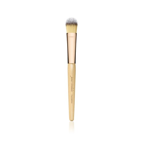 Na zdjęciu znajduje się pędzel do makijażu marki Jane Iredale. Ma elegancki design – drewniany trzonek w naturalnym kolorze z złotą skuwką i gęste, miękkie włosie w odcieniach żółci i bieli.