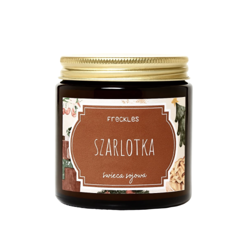 Na zdjęciu znajduje się świeca sojowa marki Freckles o zapachu „Szarlotka”. Produkt umieszczony jest w szklanym, ciemnym słoiczku z ozdobną etykietą w stylu rustykalnym i złotą zakrętką. Etykieta przedstawia nazwę zapachu