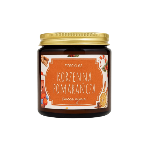 Na zdjęciu znajduje się świeca sojowa marki Freckles o nazwie Korzenna Pomarańcza. Produkt umieszczony jest w szklanym słoiczku z metalową, złotą zakrętką. Etykieta w odcieniu pomarańczowym ozdobiona jest świątecznymi grafikami przypraw