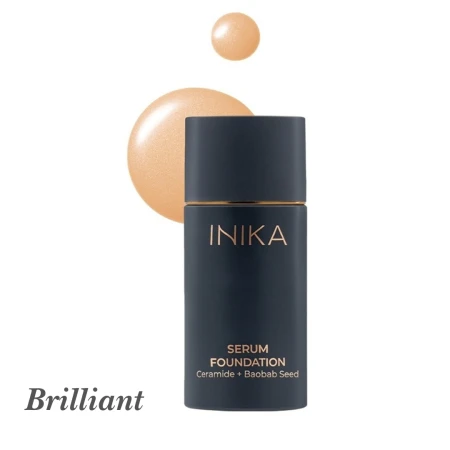 Na zdjęciu przedstawiono odcień Brilliant z gamy INIKA Organic Serum Foundation. Produkt znajduje się w eleganckiej buteleczce w matowym, granatowym kolorze ze złotymi akcentami. Obok opakowania widoczna jest próbka koloru
