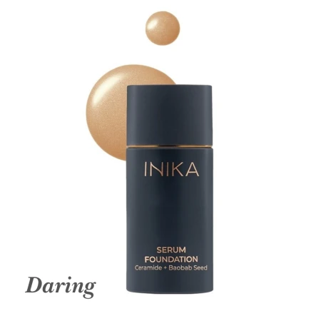 Na zdjęciu widoczny jest odcień Daring z linii INIKA Serum Foundation. Produkt jest w eleganckiej, granatowej butelce z matowym wykończeniem i złotymi detalami. Obok opakowania znajduje się próbka koloru – średni beż o neutralnym, lekko ciepłym podtonie