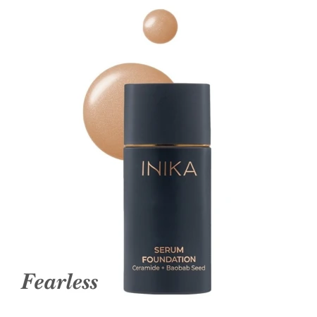 Na zdjęciu przedstawiono odcień Fearless z kolekcji INIKA Organic Serum Foundation. Produkt jest w  granatowej butelce z matowym wykończeniem i złotym akcentem. Obok znajduje się próbka koloru – ciepły, średni do ciemniejszego beż z delikatnym blaskiem.