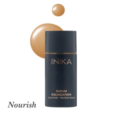 Na zdjęciu znajduje się podkład INIKA Organic Serum Foundation w odcieniu Nourish. Produkt jest w eleganckiej, matowej granatowej butelce  ze złotym napisem. Obok pokazano próbkę koloru – ciepły, średni odcień beżu z delikatnym świetlistym połyskiem.