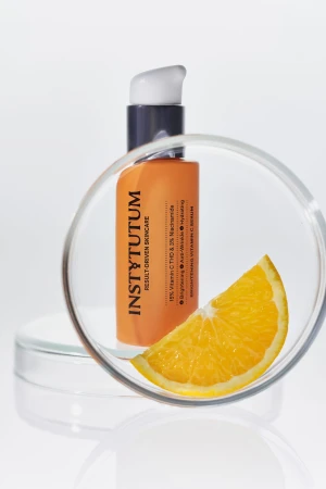 Na zdjęciu znajduje się produkt marki Instytutum Brightening Vitamin C Serum w eleganckiej, pomarańczowej butelce z pompką oraz kawałek pomarańczy