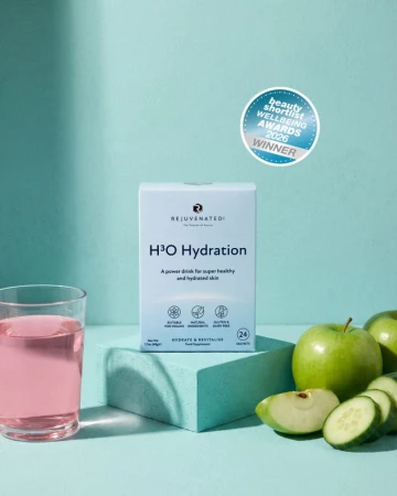 Białe opakowanie H3O Hydration marki Rejuvenated na turkusowym tle. Obok szklanka różowego napoju, zielone jabłko i ogórek. Widoczna plakietka nagrody Beauty Wellbeing Awards 2026 Winner.jpg