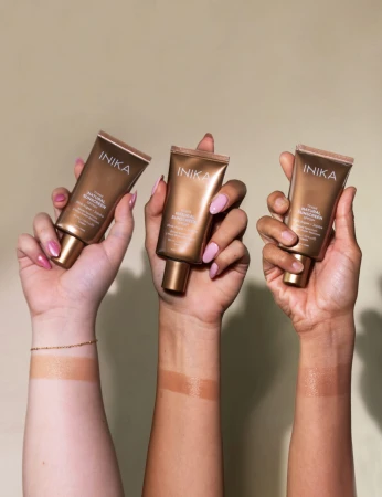 Na zdjęciu trzy osoby trzymają w dłoniach tuby INIKA Tinted Natural Sunscreen SPF50+. Widać różne odcienie skóry oraz paski produktu na przedramionach, pokazujące efekt koloru i dopasowanie do różnych karnacji.