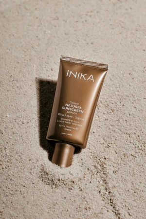 Na zdjęciu widoczna jest tuba INIKA Tinted Natural Sunscreen SPF50+ leżąca na jasnym piasku. Opakowanie ma brązowo-złoty kolor z białym logo i informacją o filtrze SPF50+, Pink Algae + Jojoba oraz 2-godzinnej wodoodporności.