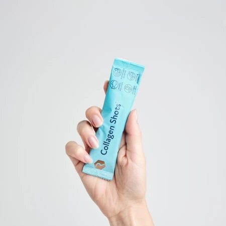 Zdjęcie przedstawia dłoń trzymającą pojedynczą, jasnoniebieską saszetkę suplementu diety w proszku Rejuvenated Collagen Shots. Produkt ten jest przeznaczony do wspierania jędrności, elastyczności i nawilżenia skóry.