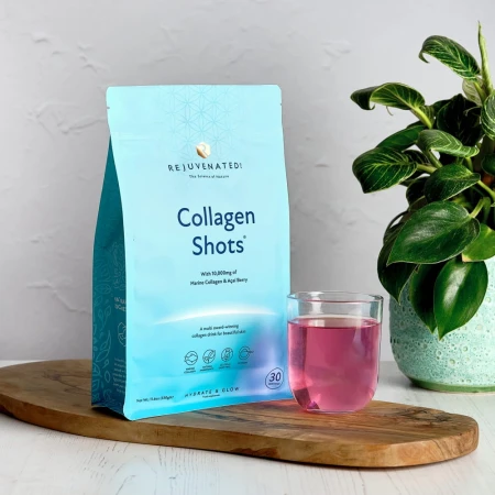 Zdjęcie przedstawia jasnoniebieskie opakowanie suplementu diety Rejuvenated Collagen Shots i szklankę różowego napoju na drewnianej desce. Obok stoi roślina doniczkowa.