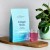 Zdjęcie przedstawia jasnoniebieskie opakowanie suplementu diety Rejuvenated Collagen Shots i szklankę różowego napoju na drewnianej desce. Obok stoi roślina doniczkowa.