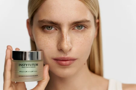 Modelka trzyma w dłoni słoiczek Instytutum Superbiotic Regeneration Cream. Krem przedstawiony jest w eleganckim, matowym opakowaniu z metaliczną nakrętką. Ujęcie podkreśla efekt zdrowej, gładkiej i promiennej skóry