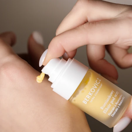 Na zdjęciu widoczna jest aplikacja Rumiankowego Eliksiru pod oczy Berkovicz Tatra Cosmetics. Jedna pompka produktu w złocistym odcieniu zostaje wyciśnięta na skórę, prezentując lekką, kremowo-żelową konsystencję.