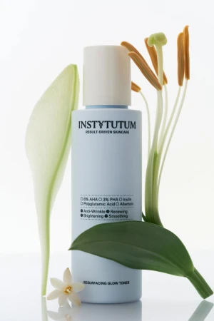 Na zdjęciu widać butelkę toniku INSTYTUTUM Resurfacing Glow Toner w jasnoniebieskim opakowaniu, ustawioną wśród zielonych liści i kwiatów na jasnym tle.