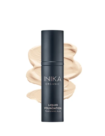 Na zdjęciu widoczny jest podkład INIKA Organic Liquid Foundation z kwasem hialuronowym w eleganckim, ciemnym opakowaniu, zaprezentowany na tle rozprowadzonego produktu, który podkreśla jego kremową konsystencję i naturalne wykończenie makijażu.