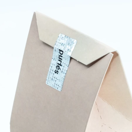 Grafika przedstawia tylne zamknięcie opakowania Purlés Cosmetic Headband. Kartonowe pudełko jest zabezpieczone srebrną naklejką z logo marki Purlés.