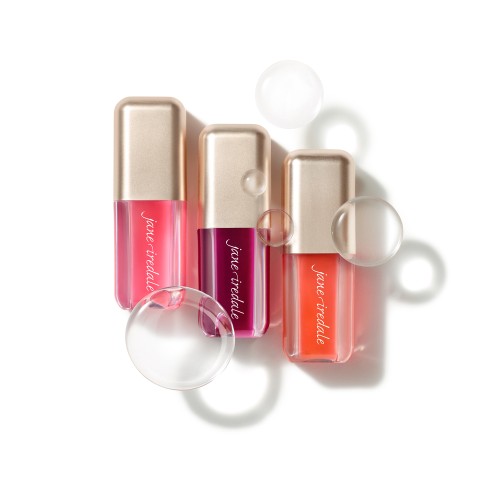Na białym tle są trzy olejki do ust Jane Iredale Just Kissed Lip Oil w przezroczystych opakowaniach z metalicznymi, złotymi nakrętkami. Olejki mają odcienie intensywnego różu, jagodowego fioletu i koralu.