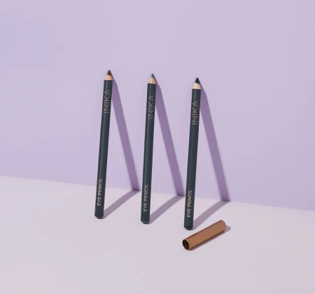 Na zdjęciu widoczne są trzy kredki do oczu ustawione pionowo na jasnym tle z lekkim fioletowym odcieniem. Każda kredka ma ciemny korpus z napisem „INIKA EYE PENCIL”, a ich końcówki różnią się kolorami. Obok leży złota skuwka.