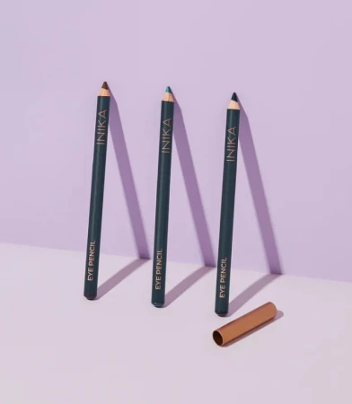 Na zdjęciu widoczne są trzy kredki do oczu ustawione pionowo na jasnym tle z lekkim fioletowym odcieniem. Każda kredka ma ciemny korpus z napisem „INIKA EYE PENCIL”, a ich końcówki różnią się kolorami. Obok leży złota skuwka.